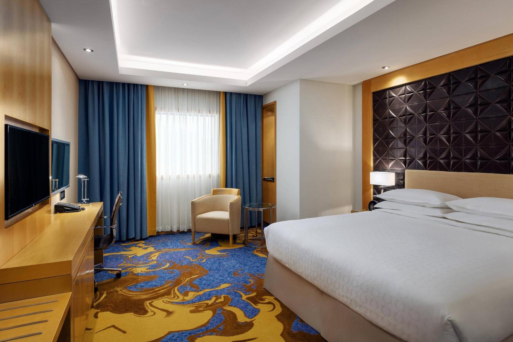 מכה Sheraton Makkah Jabal Al Kaaba Hotel מראה חיצוני תמונה The photo shows a modern hotel room featuring a comfortable double bed with a light-colored headboard. There's a large window dressed with blue curtains, allowing natural light to enter. The walls are painted in neutral tones, and one wall has a text