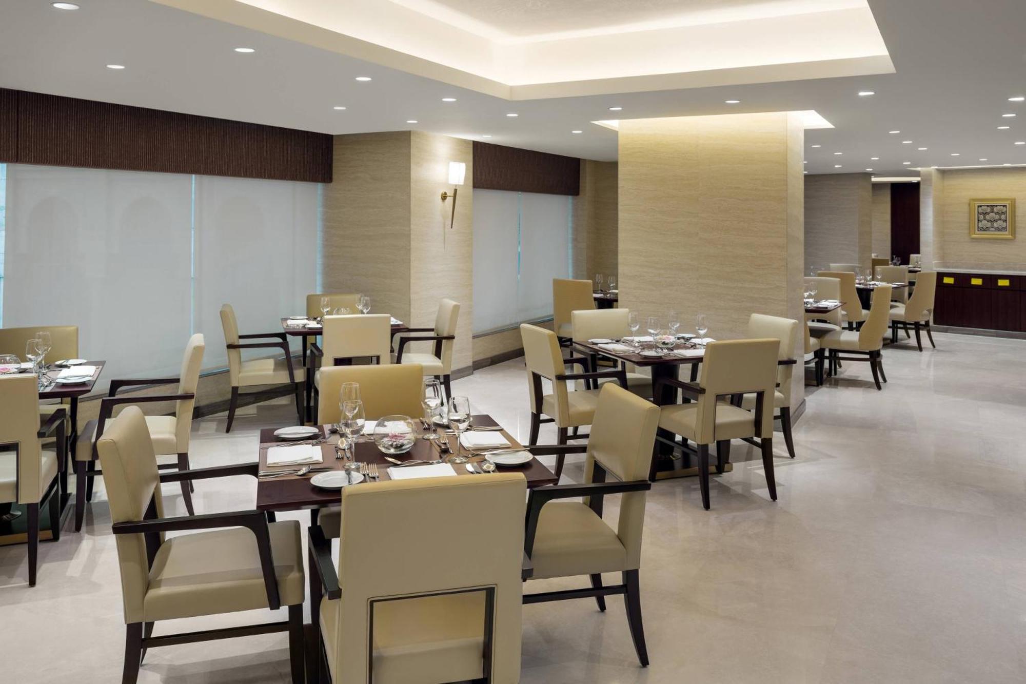 מכה Sheraton Makkah Jabal Al Kaaba Hotel מראה חיצוני תמונה The photo shows a modern restaurant interior. It features several dining tables, each set with utensils and tableware. The chairs are upholstered in a light fabric, and the overall decor is sleek and minimalistic, with soft lighting from overhead fix